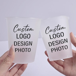 May include: Two frosted plastic cups with the words "Custom LOGO DESIGN PHOTO" printed in black. The cups are held by hands, suggesting a promotional or personalized product. The cups are cylindrical with a slightly flared top.