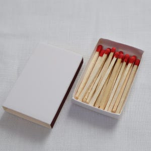 May include: A rectangular white matchbox with a sliding cover placed beside an open tray containing a collection of wooden matches. Each match features a light tan wooden stem and a distinct, rounded red tip, arranged neatly within the box.