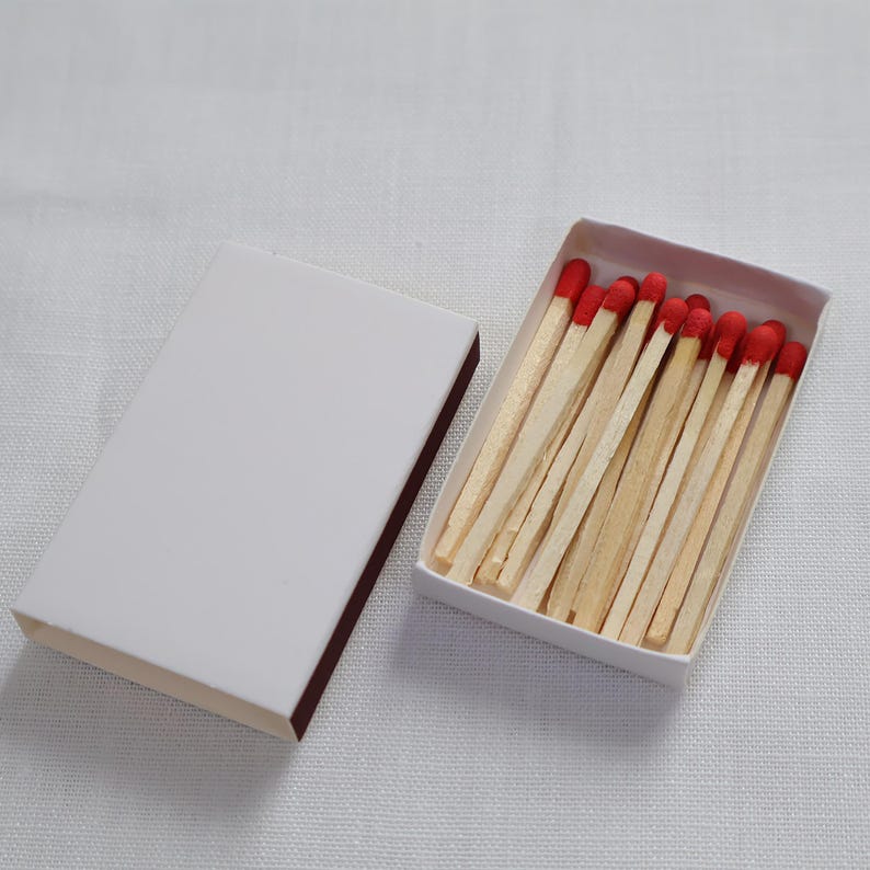May include: A rectangular white matchbox with a sliding cover placed beside an open tray containing a collection of wooden matches. Each match features a light tan wooden stem and a distinct, rounded red tip, arranged neatly within the box.