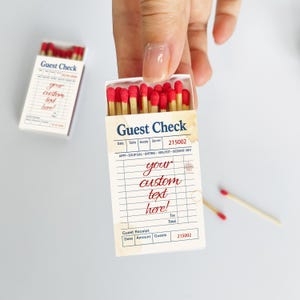 May include: A matchbox with a "Guest Check" design, filled with red-tipped matches. The box features a vintage-style receipt layout with the words "your custom text here!" printed in red. Two loose matches are visible.