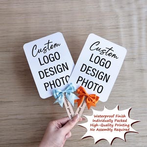 May include: Two white rectangular signs with the text "Custom LOGO DESIGN PHOTO" in black. One sign has a light blue bow, the other an orange bow. The signs are held by wooden sticks. Text on the sticks reads "custom your text".