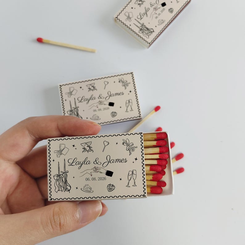 May include: Rectangular matchboxes with a cream-colored exterior featuring black line art of wedding icons, the names Layla & James, and the date 06.08.2026. The boxes contain wooden matches with bright red tips, arranged in a row inside the open box.