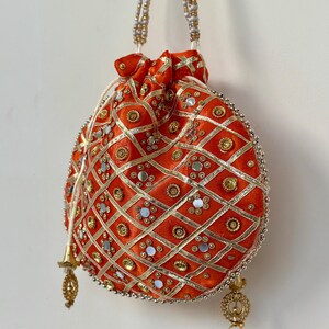 Könnte beinhalten: Ein orangefarbener Beutel mit Kordelzug und goldenen und silbernen Verzierungen. Die Tasche hat ein Rautenmuster mit goldenen Zierleisten und kleinen Spiegeln. Der Griff besteht aus weißen und goldenen Perlen, und die Tasche hat goldene Quasten.