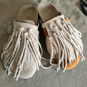 May include: A pair of beige suede sandals with long fringe detailing. The sandals have a buckle closure and a cork footbed. The soles are a light brown colour. The sandals are handmade and appear to be new.