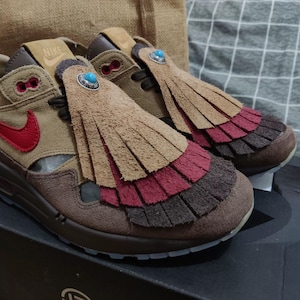 May include: Tan and brown sneakers with red accents and a fringed tongue. The shoes feature a red Nike swoosh and a turquoise stone detail. The soles are brown and the shoes are on a black box.
