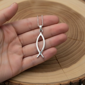 Puede incluir: Un collar con colgante plateado con el símbolo Ichthys, una forma de pez. El colgante cuelga de una fina cadena. El collar se muestra en una mano, sobre un fondo de madera con vetas circulares.