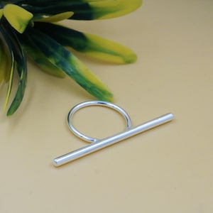 May include: A minimalist silver ring with a straight bar design. The ring has a circular band connected to a horizontal bar, creating a modern and geometric aesthetic. The ring is set against a neutral background with green and yellow leaves.