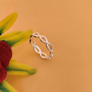 May include: A silver ring featuring an infinity symbol pattern. The ring sits on a yellow and orange surface, with a flower in the corner. This delicate piece of jewellery is a stylish accessory.