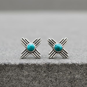 Op de afbeelding: Een paar zilverkleurige oorstekers. Elke oorbel heeft een turquoise steen in het midden van een kruisvormig ontwerp. Het kruisontwerp heeft meerdere parallelle lijnen die zich naar buiten uitstrekken.