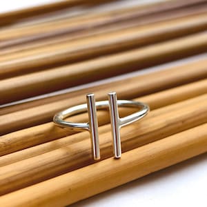 May include: A silver ring with two vertical bars. The ring is set against a background of parallel wooden rods. The ring's design is minimalist and modern, with a polished finish.