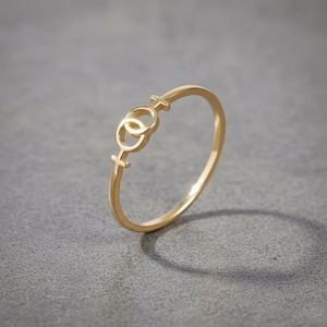 May include: A gold-colored ring featuring a double female symbol design. The ring has a thin band and a delicate, openwork design. The ring is set against a gray background.