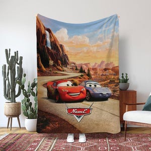 May include: A large, rectangular blanket featuring a scene from the animated movie "Cars." The design showcases Lightning McQueen and Sally Carrera on a desert road with a scenic background of mountains and Route 66 signage. The blanket is primarily in shades of red, blue, and brown.