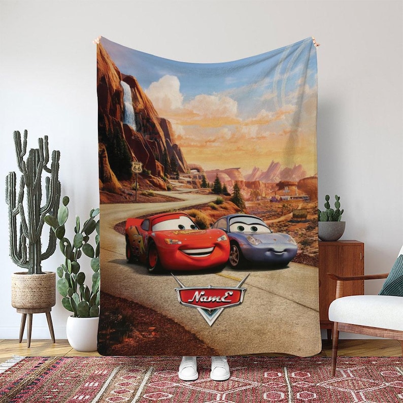 May include: A large, rectangular blanket featuring a scene from the animated movie "Cars." The design showcases Lightning McQueen and Sally Carrera on a desert road with a scenic background of mountains and Route 66 signage. The blanket is primarily in shades of red, blue, and brown.