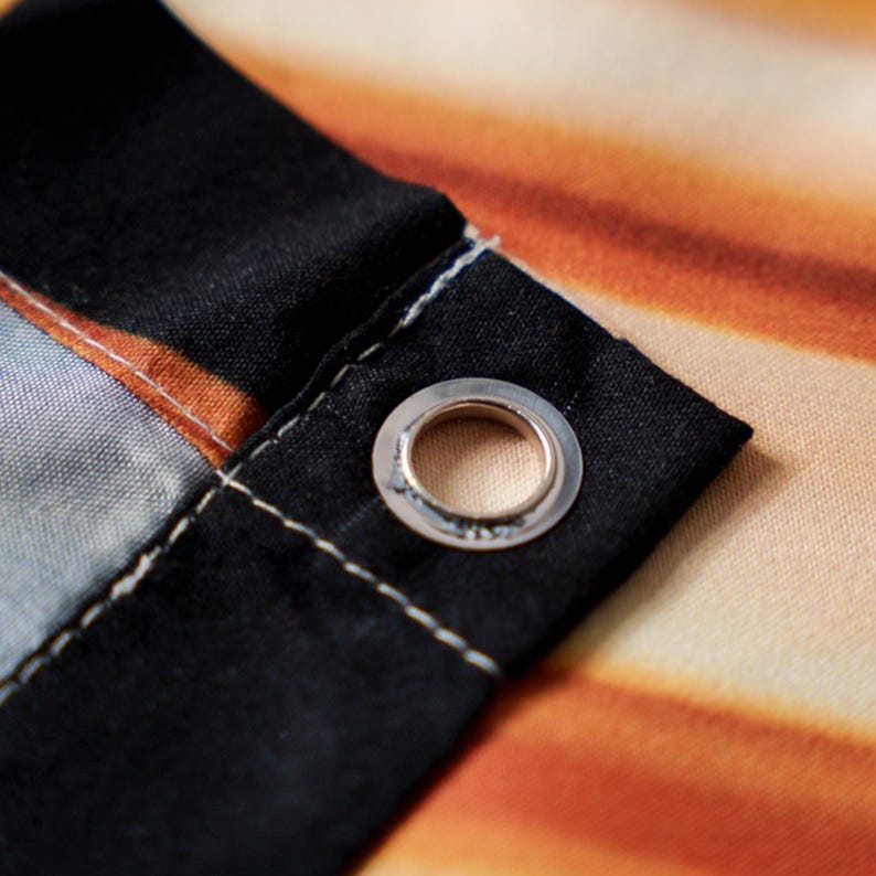May include: Close-up of a fabric edge with a metal grommet. The fabric features a gradient of orange, yellow, and black colors. The grommet is silver and reinforced with white stitching.