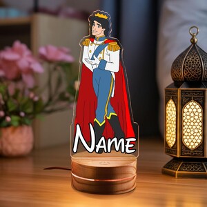May include: A decorative night light featuring a cartoon prince in a white and blue outfit with a red cape and gold crown. The base is wood with a copper accent, and the word "Name" is written on the front.