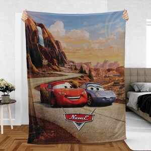 May include: A plush blanket featuring a scenic Route 66 landscape with two cartoon cars, a red one and a blue one, on a winding road. The blanket has a logo that says "Name" in a stylized font.