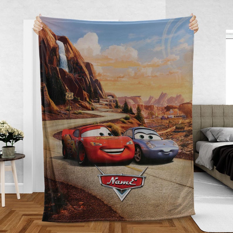 May include: A plush blanket featuring a scenic Route 66 landscape with two cartoon cars, a red one and a blue one, on a winding road. The blanket has a logo that says "Name" in a stylized font.