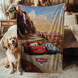 May include: A plush blanket featuring a scenic desert landscape with a waterfall and Route 66 sign. The blanket showcases the animated characters Lightning McQueen and Sally Carrera from the movie Cars. The blanket has the name "Name" on it.