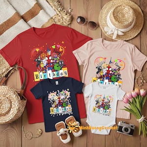 Puede incluir: Una colecci&oacute;n de camisetas rojas, azul marino y rosa claro, y un body blanco, todos con un gr&aacute;fico colorido con personajes de dibujos animados, la palabra "Birthday" y el a&ntilde;o "2026". Las prendas est&aacute;n dispuestas sobre una superficie de madera con accesorios.