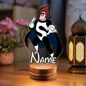 May include: A decorative night light featuring a cartoon superhero character with red hair, a black and white costume, and a blue cape. The base is made of wood and has a warm glow. The word "Name" is written in a script font.