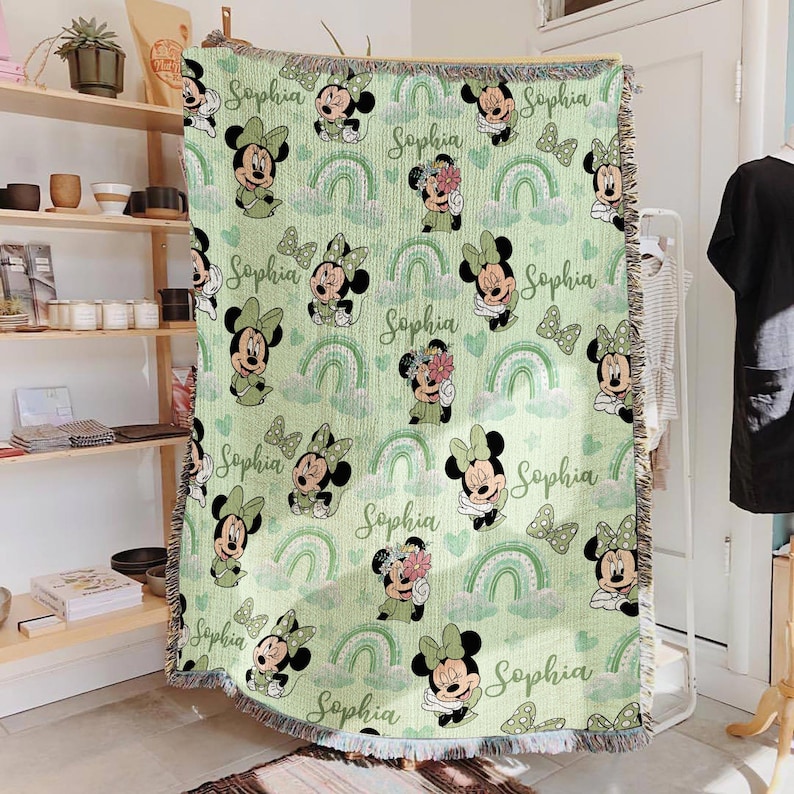 May include: A light green throw blanket featuring a repeating pattern of Minnie Mouse, rainbows, and the name "Sophia" in a script font. The blanket has a fringed edge and is made of a soft material.