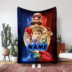 May include: A large, rectangular blanket featuring characters from the Sonic the Hedgehog movie. The design includes a central circular image with a boy's face and the word "NAME" in blue. The background is a mix of red and blue.