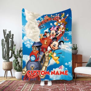 Könnte beinhalten: Eine blaue Decke mit einer farbenfrohen Illustration von Mickey Mouse, Minnie Mouse und anderen Disney-Figuren auf einem Zug. Die Decke trägt den Text "CUSTOM NAME" in roten Buchstaben.