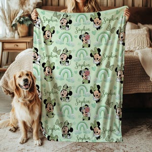 May include: A light green blanket featuring a pattern of Minnie Mouse illustrations, rainbows, and butterflies. The name "Sophia" is written in cursive throughout the design. The blanket is held up by a person.