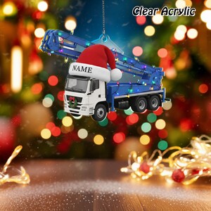 May include: Clear acrylic Christmas ornament shaped like a white and blue concrete pump truck, decorated with string lights and a Santa hat. The hat has the word "NAME" on it. A small wreath is on the front of the truck.