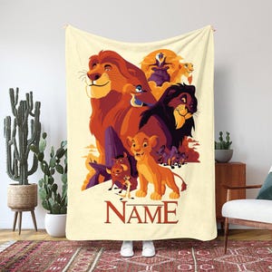May include: A cream-colored blanket featuring a colorful illustration of characters from The Lion King. The design includes Simba, Mufasa, Scar, Timon, Pumbaa, and Zazu. The word "NAME" is printed in a dark red font at the bottom of the blanket.