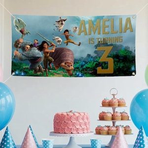 May include: A banner with the text "AMELIA IS TURNING 3" in gold lettering, featuring animated characters. The scene includes a pink cake, cupcakes, party hats, and balloons, creating a festive birthday setting.