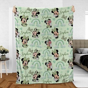 May include: A light green blanket featuring a repeating pattern of Minnie Mouse, bows, rainbows, clouds, and hearts. The name "Sophia" is written in cursive throughout the design. The blanket is held up, showcasing the full pattern.