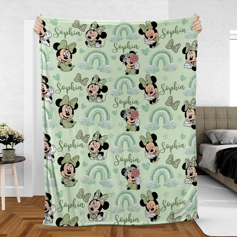 May include: A light green blanket featuring a repeating pattern of Minnie Mouse, bows, rainbows, clouds, and hearts. The name "Sophia" is written in cursive throughout the design. The blanket is held up, showcasing the full pattern.