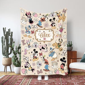 Puede incluir: Manta beige con un estampado de personajes de Disney, incluyendo Mickey Mouse, Minnie Mouse, Donald Duck y Goofy. La manta tiene un nombre personalizado, "Alex", en un marco decorativo, y está decorada con flores, arcoíris y fresas.