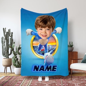 May include: A blue fleece blanket with a child's face integrated into a cartoon character design. The child is in a blue outfit with white gloves and shoes, emerging from a golden ring. The word "NAME" is displayed in blue neon text.