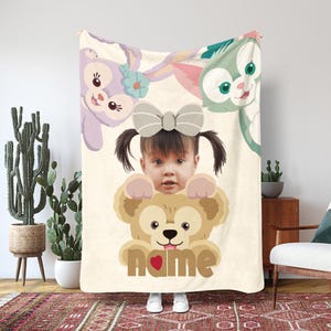 May include: A personalized cream-colored blanket featuring a child's face and the name "name" in brown letters. The blanket has cartoon characters and a large bow. The child is holding a brown bear.