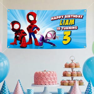 Op de afbeelding: Een blauwe banner met Spider-Man-personages en de tekst "HAPPY BIRTHDAY LIAM IS TURNING 3". De banner is versierd met een blauw stralenpatroon. Feestdecoraties zijn onder meer een roze taart, cupcakes en ballonnen.