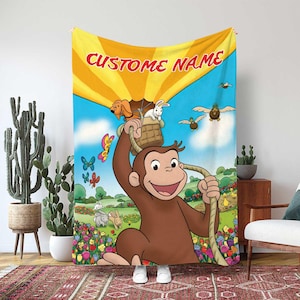 May include: A colorful blanket featuring a cartoon illustration of a monkey holding a rope attached to a hot air balloon. The blanket has the text "CUSTOM NAME" on the top. The background includes butterflies, flowers, and other cartoon animals.