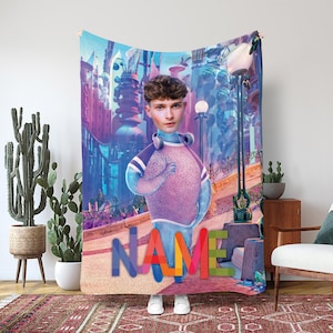 May include: A tapestry featuring a digitally created image of a person with a blue body and a purple shirt, standing in front of a colorful cityscape. The word "NAME" is displayed in large, multicolored letters at the bottom of the tapestry.