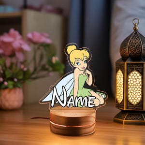 May include: A decorative night light featuring a cartoon fairy in a green dress and wings, with the word "NAME" in white script. The light sits on a wooden base with a warm glow, and a power cord extends from the base.
