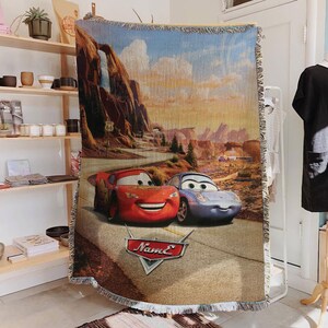 May include: Woven throw blanket featuring Lightning McQueen and Sally Carrera from the movie Cars, set against a desert landscape with Route 66 signage. The blanket has a beige fringe and a logo that says "Name E".