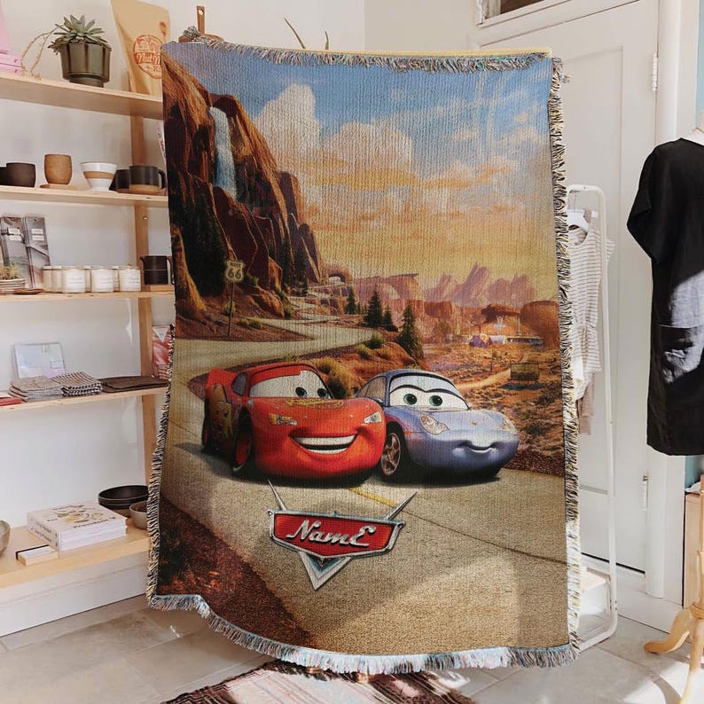 May include: Woven throw blanket featuring Lightning McQueen and Sally Carrera from the movie Cars, set against a desert landscape with Route 66 signage. The blanket has a beige fringe and a logo that says "Name E".