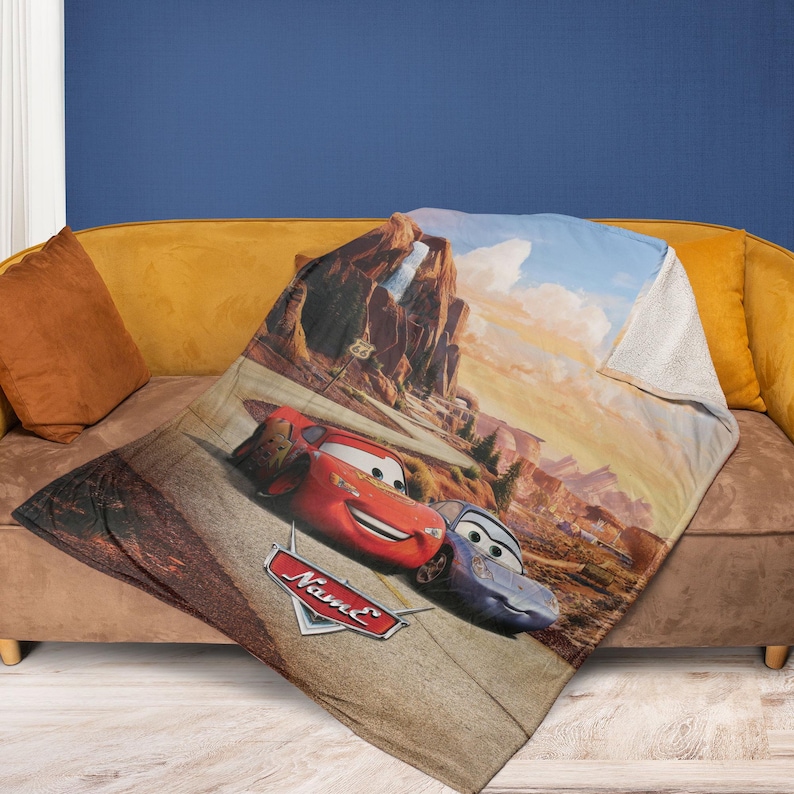 May include: A plush blanket featuring Lightning McQueen and Sally Carrera from the Cars movie. The blanket showcases a desert landscape with Route 66 signage. The Cars logo is visible on the blanket.