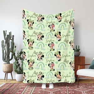 May include: A light green blanket featuring a repeating pattern of Minnie Mouse in various poses, green bows, rainbows, and the name "Sophia" in script. The blanket has a soft, pastel color scheme and is likely made of a cozy material.