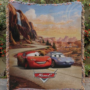May include: Woven throw blanket featuring Lightning McQueen and Sally Carrera from the movie Cars, set against a desert landscape with Route 66 signage. The blanket has a fringed edge and the word "Name" on the bottom.
