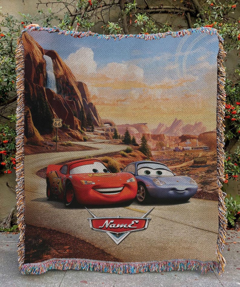 May include: Woven throw blanket featuring Lightning McQueen and Sally Carrera from the movie Cars, set against a desert landscape with Route 66 signage. The blanket has a fringed edge and the word "Name" on the bottom.