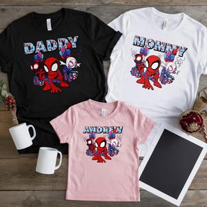 Puede incluir: Tres camisetas: negra, blanca y rosa. Cada una presenta un gr&aacute;fico de Spider-Man con los textos: "DADDY", "MOMMY" y "ANDREW". Los dise&ntilde;os incluyen globos y cajas de regalo. Se ven dos tazas blancas.