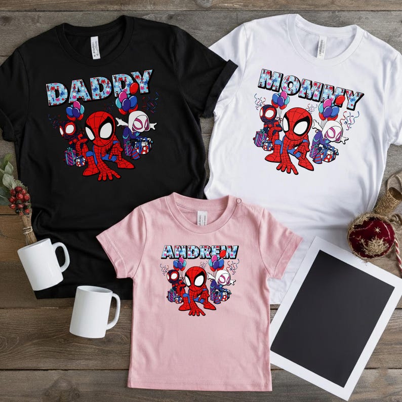 Puede incluir: Tres camisetas: negra, blanca y rosa. Cada una presenta un gr&aacute;fico de Spider-Man con los textos: "DADDY", "MOMMY" y "ANDREW". Los dise&ntilde;os incluyen globos y cajas de regalo. Se ven dos tazas blancas.