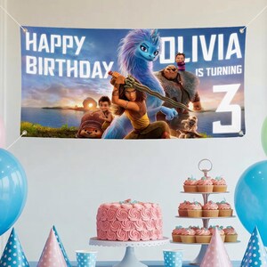 May include: A banner with the text "HAPPY BIRTHDAY OLIVIA IS TURNING 3" featuring characters from a movie. The banner is blue and white. A pink cake and cupcakes are on a table with blue and pink balloons.