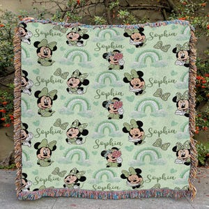May include: A light green throw blanket featuring a repeating pattern of Minnie Mouse, rainbows, and butterflies. The name "Sophia" is written in cursive throughout the design. The blanket has a fringed edge.